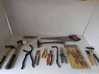 Photo showing the collection of 14 hand tools arranged on a white surface, including 3 hammers, mallet, locking pliers, handsaw, various pliers and cutters.