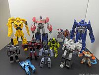 Lot of 11 Transformer robot toys arranged in two rows on a flat surface showing various characters such as yellow Bumblebee-like figure, red Optimus Prime-type, blue canine figure, and others