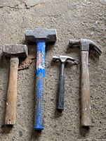 Four hammers of varying size and condition are displayed on a rough surface, showing clear use wear on wooden, blue plastic, and rubber handles as well as metal heads.