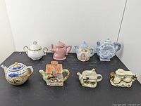 View of all 7 teapots and the covered sugar bowl arranged side by side showing variety of designs and colors.