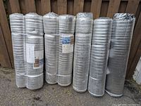 Seven rolls of silver-gray laminate flooring underlayment standing upright outside against a wooden fence; six rolls are sealed, one roll is open.