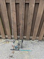 Four fishing rods leaning against a wooden fence outdoors on pavement, showing rod handles and attached reels.