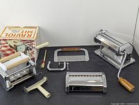 Overview of entire lot showing all pasta making tools, including the pasta roller, ravioli cutter with box, pastry cutter, pasta cutting roller, rolling pin, and brush.