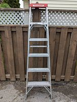 Full view of a preowned Mastercraft aluminum step ladder with five steps and a red Mastercraft label on top, photographed in front of a wooden fence outdoors.