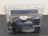 Front view of 12V air compressor in clear plastic clamshell packaging showing the compressor, gauge, power plug and hose attachments.