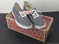 Side view of the grey Vans low top tennis shoes on top of Vans branded box, showing the shoes with white laces and tags attached.