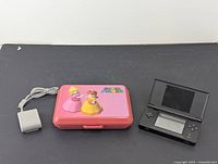Nintendo DS console open with screens and control buttons visible, pink carrying case and power adapter beside it.