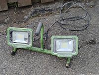 Front view of PowerSmith dual head LED work light showing green metal frame and two LED heads with attached power cable on outdoor ground surface.
