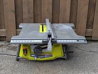 Front view of the Ryobi table saw showing gray work surface, central saw blade slot, and yellow base with control switches and adjustment knob.