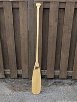 Full view of the tall, light wood-finished oar placed vertically against a dark wooden fence, showing the overall shape and condition.