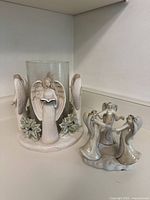 Ceramic tea light holder with four angels around a glass cylinder and holly decoration, placed on a white shelf alongside smaller candle holder with three angels.