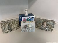 Three angel-themed block candles and three angel sculptures arranged on the floor with some small figurines, showing carved angel designs in various poses.