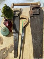 Full view of three hand saws, two oil cans, and set of three new funnels on wooden surface