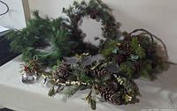 Lot of artificial greenery Christmas decorations including garland, wreath, basket, centerpiece, and hanging ornament on table.