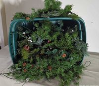 Large green plastic tote container filled with numerous artificial pine greenery sprigs with attached pinecones displayed spread out
