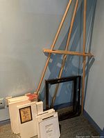 Two wooden art easels and a collection of blank canvases stacked against a wall including canvas boards and some stretched canvases.