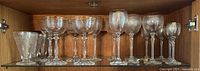 Wide view of all assorted drinking glasses on a glass shelf inside a wooden cabinet.