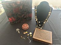 Photo showing the decorative fabric-covered box, wooden jewelry box, multi-strand necklace on display stand, small round trinket box, two tie pins and vintage buttons arranged on table.