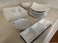 Photo showing stacked white square dinner plates, curved square plates with three branded Stokes and one unbranded, two white bowls behind the stacks, and long white rectangular dish with three compartments in front.