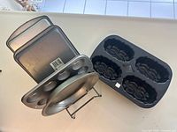 Various metal pans and molds including square cake pans, mini muffin pan, pie plate, decorative cake mold, and rack arranged on a flat surface.