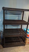 Photo showing full view of the black plastic storage shelf with four grid-style shelves supported by plastic tube legs.