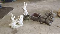 Set of four resin bunny garden ornaments arranged in a line on a concrete floor indoors. The ornaments include bunnies holding a planter, a bunny sitting in a basket, and a grey bunny with a cart basket. Close-up view of each piece visible.