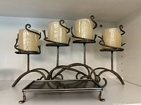 Four-tier black metal candle holder holding four cream-colored pillar candles wrapped in plastic. Scroll design and sturdy base.