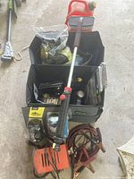 Photo showing black fabric trunk organizer filled with various automotive tools such as battery charger, jumper cables, and a wash brush with red handle, along with multiple flashlights and other small items arranged around it.