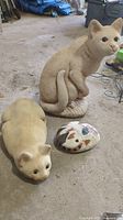 Photo showing two concrete cat garden statues and one painted rock on a garage floor, highlighting their used condition.