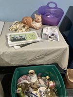 Photo showing a table with two ceramic angels, three white photo frames, a painted orange tabby cat figurine, and a small purple tote