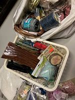 Basket containing multiple crafting materials including paper twist rolls, pipe cleaners, beads, bells, styrofoam balls, pebbles, wooden shapes, pom-poms, and stickers.