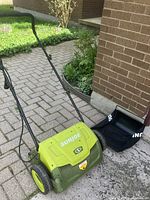Full view of the green SunJoe electric dethatcher and separate black collection bag on paved surface next to garden bed.