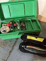 Open green fishing tackle box showing contents including fishing line spool, red and white bobbers, metal lures and swivels in separate compartments.