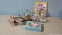 Full view of all hand painted floral home decor items on table including vases, tea light holders, trinket box, candy dishes, metal birdhouse tea light holder, and unframed floral print.