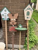 Photo showing four garden stakes and one green plastic birdbath/feeder against a brick wall and greenery background.
