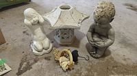 Full view of the four garden statues and decor items on concrete floor, includes the sitting child statue, garden lantern, resin child statue, and resin angel under umbrella.