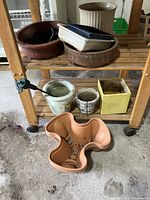 Wide view of planters on wooden rolling cart, showing various pots including round brown and cream ceramic, rectangular planter, green and white ceramic pots on lower shelf, terracotta planter on floor.