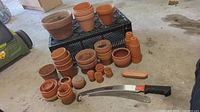 Various sizes of clay pots stacked in a plastic crate on a garage floor, with the gardening saw lying in front.