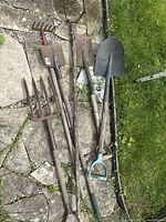 Five gardening tools laid out on stone pavement next to grass, including 2 shovels, pitchfork, garden edger, and rake.