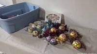 Photo showing multiple hand painted floral Christmas balls in various colors placed on a white surface next to a large gray plastic tote.