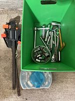 Green container holding assorted hand tools including clamps, sockets, socket wrenches, a wooden mallet, and a metal file on concrete floor, plus plastic case below.