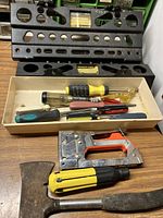 Image shows multiple screwdrivers with differently colored handles, a manual stapler, and a hatchet placed inside a white tray on a brown wooden surface with two black metal tool racks behind.