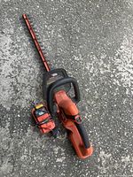 Black & Decker cordless hedge trimmer laid on pavement with attached battery and battery charger unit beside it.