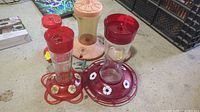 Two glass and plastic hummingbird feeders with red tops and bases, featuring white flower-shaped feeding ports, photographed on a concrete floor.