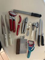 Photo showing assorted kitchen utensils including knives with white handles, red peelers, a black-handled serrated knife, ice cream scoop, and other cutting and peeling tools.