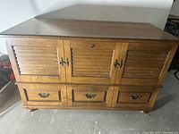 Front view of medium brown wooden cedar chest with three front compartments and metal latches. Shows surface wear and dust.