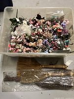 Top view of a large tote containing numerous small artificial rosette flowers in various colors, with three small twig brooms placed underneath wrapped in plastic.