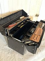 Opened black metal toolbox showing multiple compartments with assorted hand tools inside including saw, screwdrivers, chisels, and hammer.