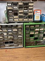 Front view of three fastener sorting containers stacked on a wooden table, showing multiple labeled drawers containing assorted nuts, bolts, screws, nails, anchors, and hardware items.