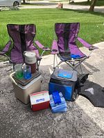 Two purple folding lawn chairs with metal frames, one with visible fabric damage, three portable coolers of various types, freezer packs and cooler accessories displayed outdoors on pavement